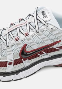 Nike-sneakers har en vit meshuvuddel med grå och vinröda accenter, en svart swoosh och en strukturerad sula. Snörningarna är grå med röda ränder.