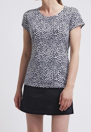 Woman wearing a short-sleeve black and white spotted top paired with a plain black mini skirt, standing against a plain background.