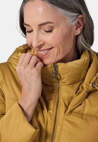 Pufferjacke in senfgelb, mit hohem Kragen, Reißverschluss vorne mit Metallzug und einer glatten, weichen Textur.