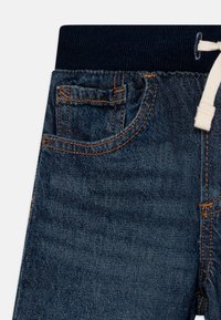 Jean en denim bleu foncé avec une taille côtelée marine. Comprend une petite poche avant et des coutures orange contrastantes. Tissu doux et texturé.