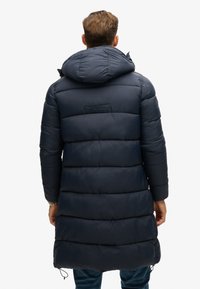 Lang, mørkeblå pufferjakke med hætte, vandret quiltet design, snoretræk i kanten og diskret branding på bagsiden.