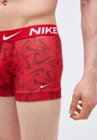 Άνω μέρος ανδρικού σώματος και χέρι με κόκκινα ανδρικά μποξεράκια Nike με λευκό λογότυπο στη μέση και στο πόδι, στέκεται μπροστά σε απλό φόντο.