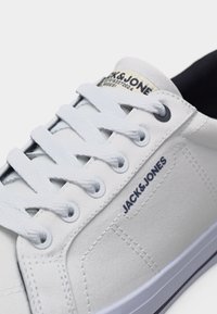 Λευκό αθλητικό παπούτσι με κορδόνια, με μαύρο λογότυπο "JACK & JONES" στο πλάι, λευκή σόλα με μαύρη ρίγα και μαύρη εσωτερική επένδυση.