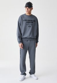 Šedá mikina a odpovídající kalhoty s nápisem "Los Angeles Lakers". Měkká tkanina, uvolněný střih, s bílými teniskami a černou šiltovkou.