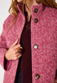 Cappotto di lana rosa con un colletto alto, caratterizzato da grandi bottoni marroni e una finitura testurizzata, indossato sopra un top marrone scuro.