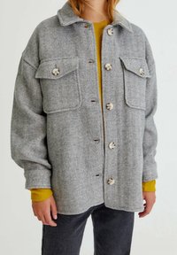 Personne portant une veste à boutons en chevrons gris avec de grandes poches avant, par-dessus une chemise jaune moutarde à manches longues et un pantalon sombre.
