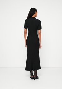 Femeie cu păr creț, purtând o rochie midi neagră mulată cu mâneci scurte și pantofi negri cu toc, stând în fața unui fundal alb.