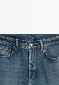 Jeans in denim blu con bottone frontale, passanti per cintura e tasche, stesi in piano con l'etichetta Massimo Dutti visibile all'interno della vita.