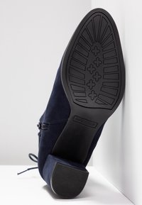 Semelle d'une bottine en daim bleu marine avec talon bloc, semelle à motifs, fermeture éclair latérale, et marque « ANNA FIELD » embossée sur la semelle.