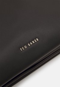 Accessorio in pelle nera con il marchio "Ted Baker" in oro impresso sulla superficie, che mostra una texture liscia e una finitura lucida.