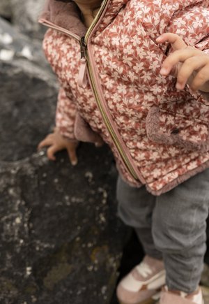 Niño pequeño con chaqueta rosa de flores, pantalones grises y zapatos rosas, de pie y tocando una superficie de piedra áspera y oscura al aire libre.