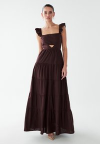 Vrouw staat en draagt een mouwloze donkerbruine maxi-jurk met meerdere lagen, gerimpelde bandjes en een kleine uitsparing aan de voorkant.