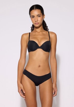 Donna con capelli ricci che indossa un reggiseno nero e biancheria intima nera coordinata, in piedi davanti a uno sfondo semplice, rivolta in avanti.
