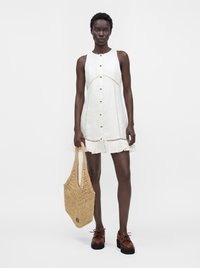 Femme portant une robe blanche sans manches boutonnée avec un ourlet à volants, tenant un grand sac fourre-tout tissé, portant des chaussures compensées marron.