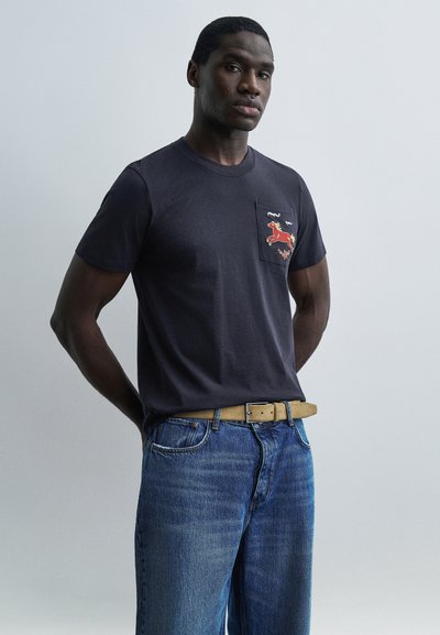 Hombre con camiseta azul marino con bordado de caballo rojo en el bolsillo del pecho, vaqueros azules y cinturón marrón claro, de pie contra un fondo claro y liso.