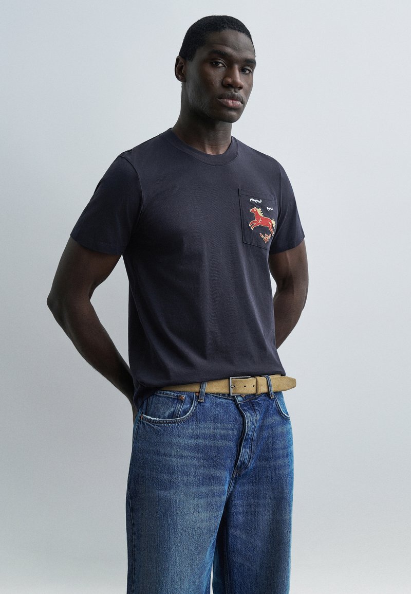 Άνδρας που φοράει μπλε ναυτικό μπλ ουζάκι με κόκκινη κεντημένη εικόνα αλόγου στην τσέπη στο στήθος, μπλε τζιν και καφετί ζώνη, στέκεται μπροστά σε απλό ανοιχτό φόντο.