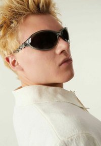 Lunettes de soleil noires avec un design élégant et incurvé et des montures grises texturées. Portées par un modèle aux cheveux blonds clairs, portant un vêtement à col blanc.