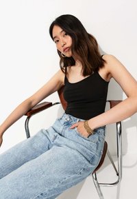 Débardeur noir ajusté avec de fines bretelles, associé à un jean en denim bleu clair taille haute. Sujet assis dans une chaise à cadre chrome.