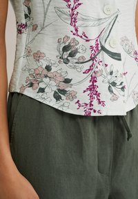 Bloemenblouse van lichte stof met roze en paarse bloempatronen, voorzien van witte knopen; gecombineerd met donkergroene linnen broek.