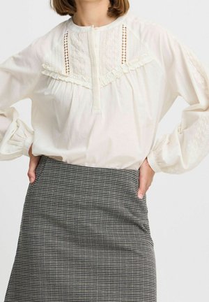 Vrouw draagt een witte blouse met lange mouwen en geborduurde details, samen met een hooggelede zwart-witte pied-de-poule rok, handen in de zij.