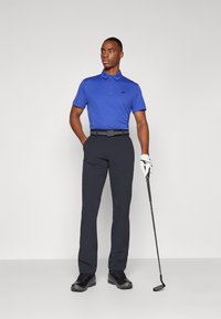 Blå kortermet poloskjorte med svart logo, kombinert med marineblå bukser og svart belte, iført svarte sko og holder en golfklubbe.