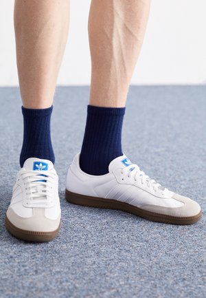 Witte leren sneakers met grijze suède accenten en een bruine rubberen zool, gecombineerd met marineblauwe geribbelde sokken, op een blauwe tapijtoppervlak.