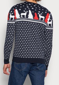 Pull bleu foncé avec motifs d'hiver blancs et rouges, comprenant des rennes et des sapins, texture tricotée, avec poignets et ourlet côtelés.