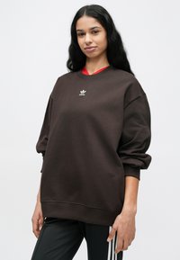 Καφέ υπερμεγέθη φούτερ με στρογγυλή λαιμόκοψη και μακριά μανίκια. Διαθέτει λευκό κεντητό λογότυπο στο στήθος. Το μοντέλο φοράει μαύρο παντελόνι με ρίγες.