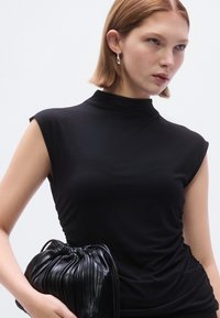 Top preto sem mangas com gola alta e laterais franzidas. Tecido liso e elástico. Segura uma mala preta de couro plissada com abertura franzida.