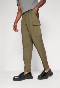 Olivgrüne Cargo-Hosen mit zwei Seitentaschen, schmal zulaufenden Beinen und einem Knopfverschluss. Kombiniert mit schwarzen Loafers mit dicken Sohlen.