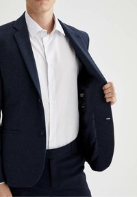Giacca da abito blu navy a trama con due bottoni, taglio slim, e foderatura nera a contrasto. Indossata sopra una camicia bianca, che mostra l'etichetta interna.