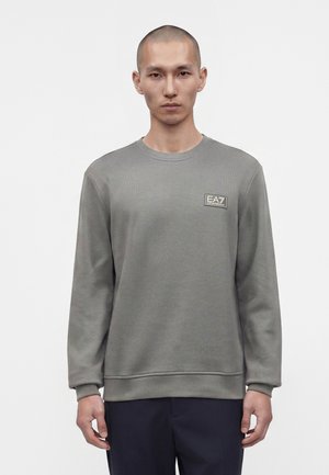 Homme portant un sweat-shirt gris uni à manches longues avec un logo EA7 sur la poitrine, debout devant un fond blanc.