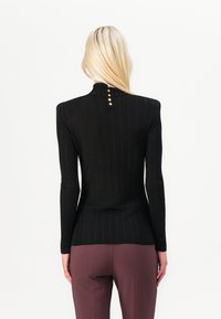 Black ribbed long-sleeve top featuring a high neck and gold-button accents at the back, paired with fitted burgundy trousers.