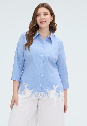 Donna che indossa una camicia a righe blu e bianca con bottoni e dettagli in pizzo floreale bianco all'orlo e pantaloni bianchi a gamba larga.