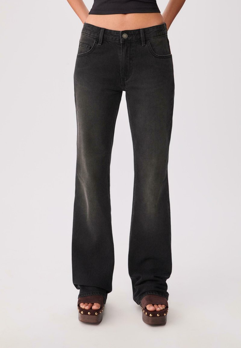 Schwarze, leicht ausgeflogene Jeans mit subtiler Ausbleichung, Fünf-Taschen-Design, Knopfverschluss und gestickten Details, kombiniert mit klobigen, spitz zulaufenden Plateau-Sandalen.