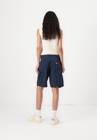 Marineblå cargo-shorts med hvite striper, med lommer, et rødt hjertepatch og en avslappet passform, sammen med hvite sneakers og sokker.