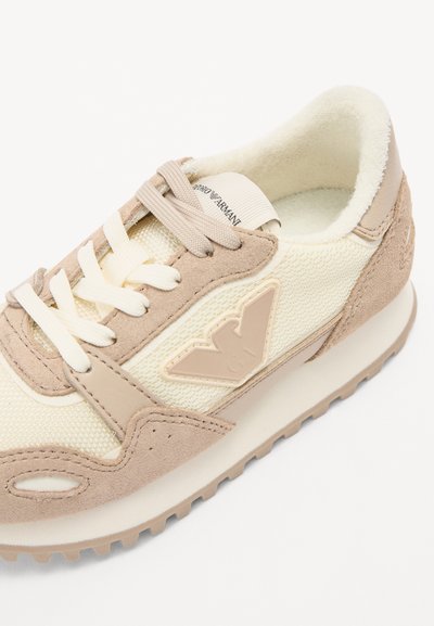 Zapatillas beige que combinan materiales de gamuza y malla, con cordones, un cuello acolchado y un logotipo embosado prominente en el lateral.