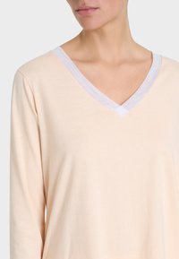 Top lungo color pesco chiaro con maniche lunghe e scollatura a V bordata di bianco. Tessuto morbido e liscio con un fit comodo.
