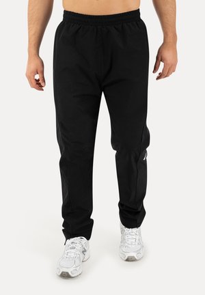 Mężczyzna w czarnych sportowych spodniach z elastycznym pasem i białych butach do biegania stoi na jednolitym tle.