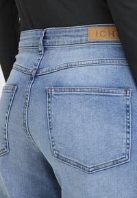 Osoba nosi svetlomodré rifle s vysokým pásom s vreckami na zadnej strane a hnedou koženou značkovou nášivkou na páse, ktoré má kombinované s čiernym topom.