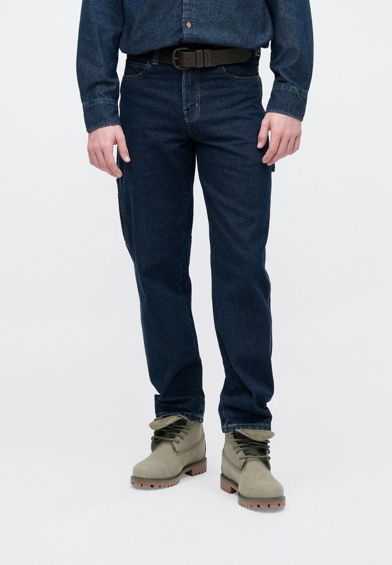 Dark blue denim jeans with a straight fit, paired with green ankle boots featuring a textured surface and brown rubber soles.