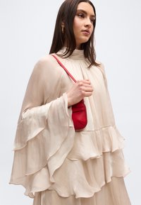 Vrouw in een vloeiende crèmekleurige jurk met geplooide mouwen, die een kleine rode schoudertas vasthoudt, tegen een lichte, effen achtergrond.