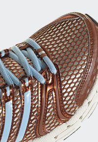 Chaussure de sport avec une tige en mesh bronze, des lacets bleu clair et des accents métalliques. Elle présente une semelle texturée et des motifs complexes sur le mesh.