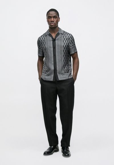 Μπλούζα με κοντά μανίκια σε μαύρο και γκρι μοτίβο γεωμετρικού σχεδιασμού, συνδυασμένη με μαύρο παντελόνι και μαύρα παπούτσια, το μοντέλο στέκεται μπροστά σε απλό φόντο.