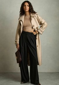 Beige trenchcoat met brede revers over een aansluitend beige top, gecombineerd met hoge zwarte geplooide broeken en een bordeauxrode handtas.