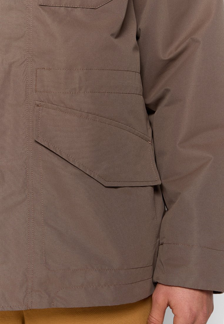 Giacca da esterno marrone con finitura testurizzata, dotata di una grande tasca laterale e cuciture a contrasto, progettata per resistenza e funzionalità.