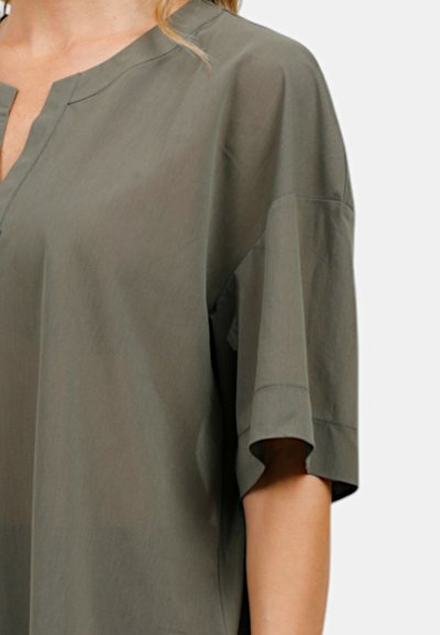 Camicetta oversize verde khaki con scollo a V, maniche corte e un tessuto liscio e leggero. Le cuciture conferiscono una sottile texture.