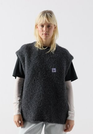 Persona bionda che indossa un maglione smanicato grigio scuro e testurizzato sopra una maglietta a maniche corte nere e una camicia bianca a maniche lunghe, in piedi contro uno sfondo bianco.