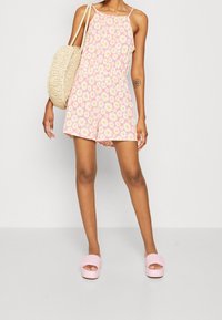 Růžový květinový overal s žlutými sedmikráskami, tenkými ramínky a elastickým pasem. Spárovaný s tkanou taškou a růžovými skluzavkami.