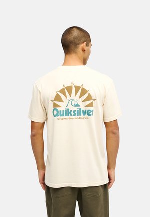 Mężczyzna z krótkimi włosami, noszący kremowy t-shirt Quiksilver z logo słońca i fali oraz napisem „Original Boardriding Co.” na plecach.